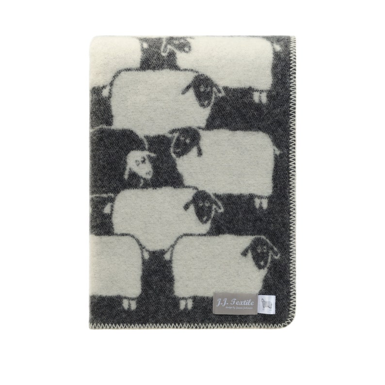 May include: A white and grey wool throw blanket with a repeating pattern of black sheep. The blanket has a woven label that reads "J-J. Textile Designed by James Johnston".