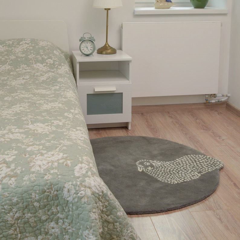 Op de afbeelding: Een rond grijs tapijt met een wit schapenontwerp in het midden. Het tapijt ligt in een slaapkamer naast een wit nachtkastje met een lamp en een klok.