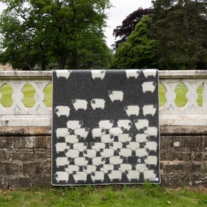 May include: A grey wool throw blanket with a white sheep pattern. The blanket is hanging on a stone fence.