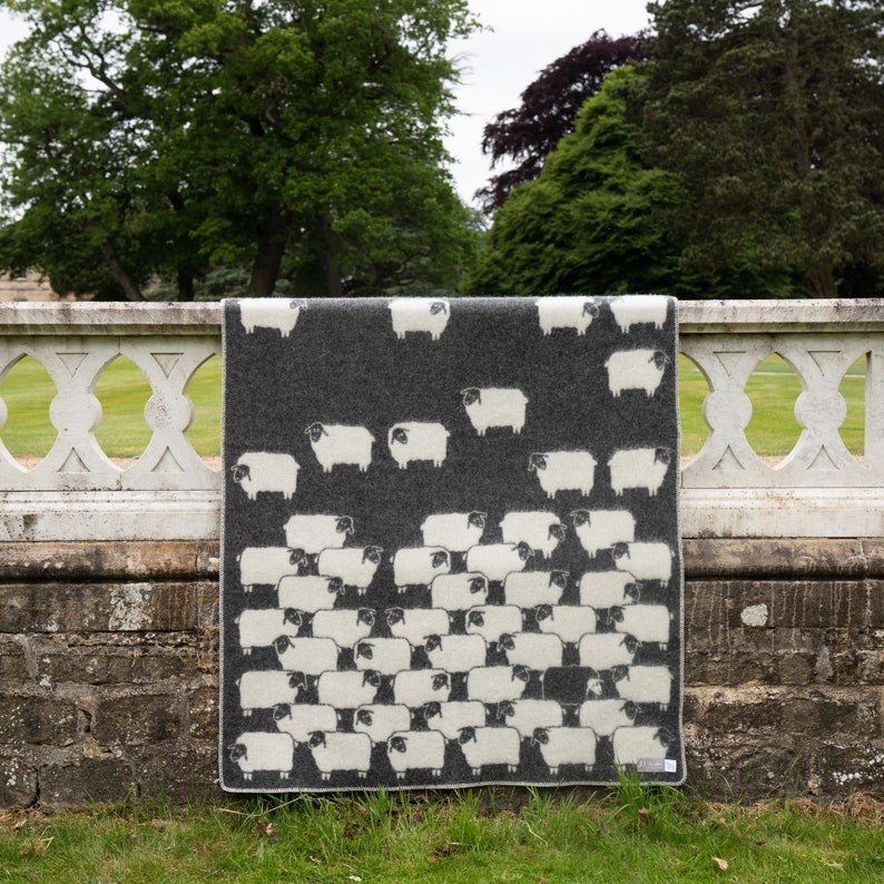 May include: A grey wool throw blanket with a white sheep pattern. The blanket is hanging on a stone fence.