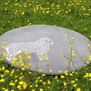 May include: Oval gray rug with a white sheep design. The rug is set in a field of green grass and yellow flowers. The sheep is depicted in a simple, dotted style.
