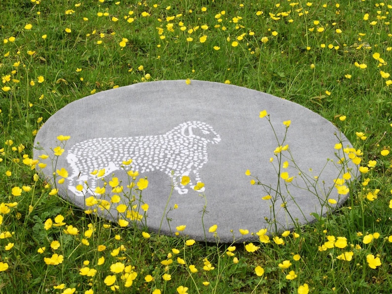 May include: Oval gray rug with a white sheep design. The rug is set in a field of green grass and yellow flowers. The sheep is depicted in a simple, dotted style.
