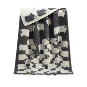 May include: A grey and white wool throw blanket with a pattern of sheep. The blanket is folded in half and has a white border.