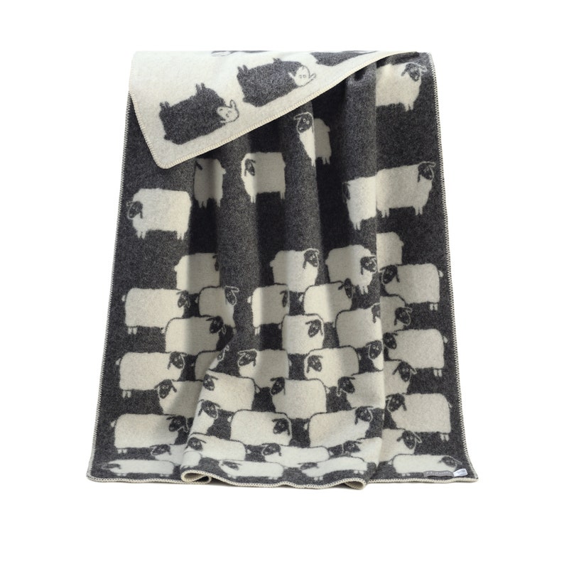 May include: A grey and white wool throw blanket with a pattern of sheep. The blanket is folded in half and has a white border.