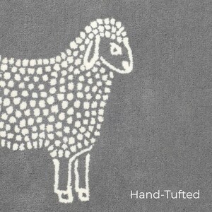 May include: A gray, hand-tufted rug featuring a white sheep design. The sheep is depicted in a simple, dotted style, with the words "Hand-Tufted" in white text at the bottom right corner.