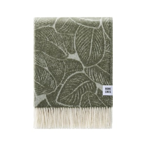 Puede incluir: Una manta de lana verde con un patrón de hojas blancas y un borde de flecos. La manta tiene una etiqueta blanca con el texto "HOME LIKES".