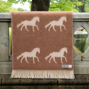 May include: A brown blanket featuring four cream-coloured horse silhouettes. The blanket has a fringed edge and a small rectangular tag in the lower right corner. The blanket is draped over a stone wall.