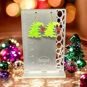 May include: A pair of green Christmas tree earrings with red glitter ornaments. The earrings are on a white display stand with a ruler and the text "Salted Suds and Creations".