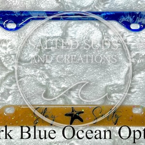 Pu&ograve; includere: Un telaio per targa a tema oceanico blu scuro e bianco con uno sfondo di glitter dorato. Il telaio ha un design a spirale bianco e blu e il testo "Stay Salty" con una stella marina.