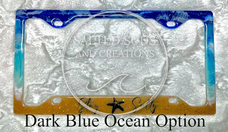 Pu&ograve; includere: Un telaio per targa a tema oceanico blu scuro e bianco con uno sfondo di glitter dorato. Il telaio ha un design a spirale bianco e blu e il testo "Stay Salty" con una stella marina.
