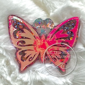 May include: Pink and iridescent butterfly-shaped resin charm with glitter embedded inside. The charm has a holographic finish and the text "SALTED SUDS AND CREATIONS" is visible on the bottom.