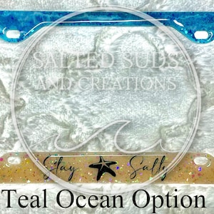 Pu&ograve; includere: Cornice per targa di colore blu-verde e bianco a tema oceanico, con glitter e una stella marina. La cornice ha il testo "Stay Salty" in basso.