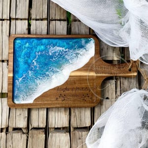 May include: Wooden serving board with a blue and white resin design resembling ocean waves. The board has a handle and engraved text that reads "Kristen & Nicholas 5/1/2015". The words "Salted Suds and Creations" are also visible.