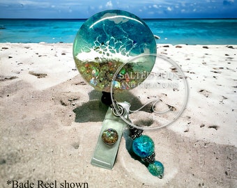 May include: A teal blue and white resin badge reel with glitter and a turquoise and silver beaded charm. The badge reel is on a white sandy beach with a turquoise ocean in the background. "Salted Goods and Creations" is written on the resin.