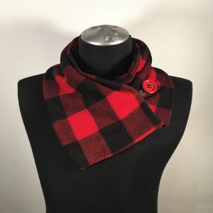 May include: A red and black buffalo plaid scarf with a large red button closure. The scarf is displayed on a black mannequin, showcasing its design and texture. The scarf is a cozy accessory.