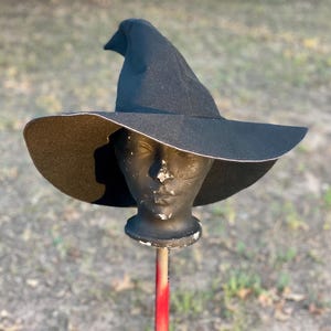 May include: A black witch hat with a wide brim and a pointed crown. The brim is trimmed with white stitching. The hat is displayed on a mannequin head mounted on a red and silver pole. The hat is made of a felt-like material.