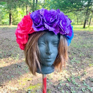 Peut inclure: Une couronne florale vibrante avec de grandes roses en tissu colorées dans les tons de rose, de violet et de bleu. La couronne est présentée sur une tête de mannequin avec des cheveux bruns, sur un fond extérieur naturel.