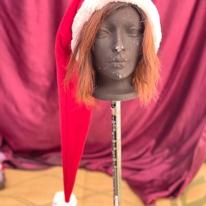 May include: A long, red Santa hat with a white faux fur trim. The hat is on a mannequin head with brown hair.