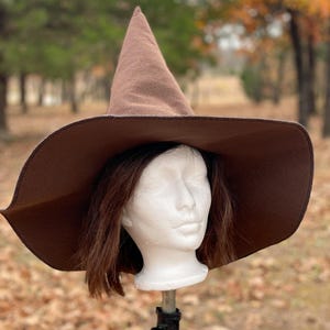Könnte beinhalten: Ein brauner Hexenhut mit breiter Krempe und spitzer Krone. Der Hut wird auf einem weißen Schaufensterkopf mit braunem Haar vor einem verschwommenen Hintergrund aus Bäumen und Laub gezeigt.