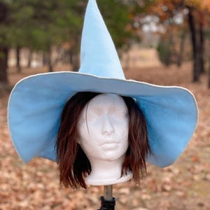 May include: A light blue witch hat with a wide brim and a pointed top. The hat is displayed on a white mannequin head with brown hair. The hat is made of a soft material and is suitable for costume parties or Halloween.