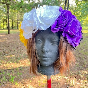 May include: A colourful floral crown with large fabric roses in white, purple, and yellow. The flowers are arranged on a headband, creating a vibrant accessory. The crown is displayed on a black mannequin head with brown hair, set against a natural outdoor backdrop.