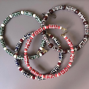 Puede incluir: Tres collares con cuentas cilíndricas que presentan patrones de rejilla y puntos blancos en verde, rojo y azul. Los diseños incluyen cuentas redondas más pequeñas en blanco, rojo y marrón oscuro, con cierres de metal dorado.