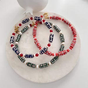 May include: Two beaded necklaces featuring cylindrical beads in red, dark blue, and dark green, each decorated with white and red circular patterns. The necklaces are accented with smaller round white and red beads and gold-toned metal clasps.