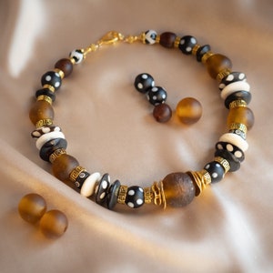 Op de afbeelding: Een handgemaakte ketting en bijpassende oorbellen set. De ketting heeft grote bruine, zwarte, witte en gouden kralen. De oorbellen zijn kleine zwarte kralen met witte stippen. De sieraden worden gepresenteerd op een satijnen oppervlak.