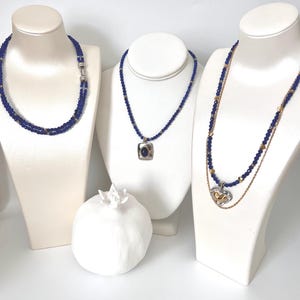 May include: Three necklaces with blue beads and gold accents are displayed on white busts. One necklace is double-stranded, another has a square pendant, and the third has a heart-shaped pendant. A white ceramic pomegranate is in the foreground.