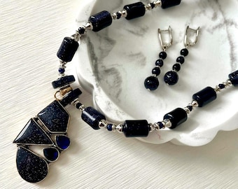 Puede incluir: Un collar y un juego de pendientes a juego de piedras preciosas azul oscuro. El collar presenta cuentas rectangulares y redondas con detalles plateados, y un colgante con formas geométricas. Los pendientes tienen un diseño colgante con cuentas redondas.