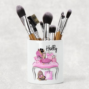 May include: White ceramic makeup brush holder with a pink and black design featuring a high heel shoe, makeup products, and a small dog. The holder has the name "Holly" written on it.