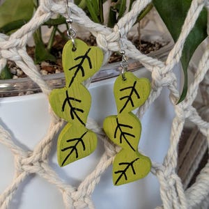 May include: A pair of dangle earrings featuring three stacked, heart-shaped lime green wooden leaves with black hand-painted vein details. The earrings hang from silver-toned metal hooks and display a visible wood grain texture on the surface.