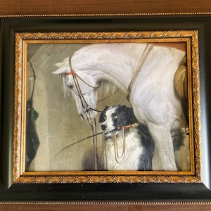 May include: Framed painting featuring a white horse and a black and white dog. The horse is saddled and bridled, and the dog holds a riding crop in its mouth. The frame is black with a gold ornate inner border. The artwork is a classic equestrian scene.