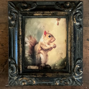 May include: A framed print featuring a detailed illustration of a squirrel holding a nut. The artwork is set within a black, ornate frame with gold accents. The squirrel is depicted in shades of brown and tan, set against a soft, muted background.
