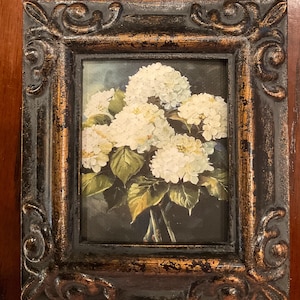 May include: A framed floral painting featuring a bouquet of white hydrangeas with green leaves. The painting is set in a rectangular, ornate black frame with gold accents, creating a vintage aesthetic.