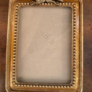 May include: A rectangular picture frame with a metallic gold finish and a beaded inner border. A sculpted metal fox figure is attached to the top center of the frame. The frame features a light beige backing insert with four small pinholes.