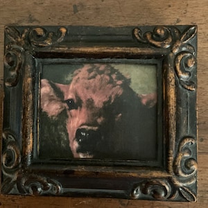 May include: A framed painting of a red cow with a dark background. The frame is black with gold accents.
