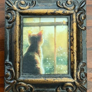May include: Small framed artwork featuring a cat looking out a window. The cat is silhouetted against a bright, yellow-toned background. The frame is ornate, with a black and gold finish, and is approximately 4 inches tall and 3 inches wide.