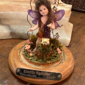 May include: A figurine of a fairy with purple wings and a brown and purple dress, standing on a wooden base with a miniature garden. The base has a metal plaque that reads "Earth Spirits".