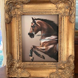 May include: A framed equestrian art print. The print features a brown horse in mid-jump, wearing a bridle and saddle. The frame is ornate, with a gold finish and detailed carvings. The artwork is displayed indoors.