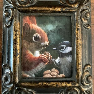 May include: A framed painting depicts a red squirrel and a small bird sharing a pine cone. The painting has a dark green background and is set in a black and gold ornate frame. A pile of nuts is at the bottom.