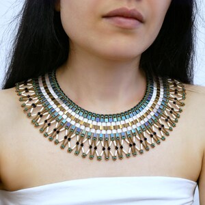 Könnte beinhalten: Ein Halsband-Halskette mit Perlenbesatz und geometrischem Design. Die Halskette hat goldene, weiße, schwarze und türkisfarbene Perlen. Die Halskette wird um den Hals getragen.