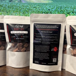 May include: Several white pouches of Buddy's Best freeze-dried pet treats are displayed. The pouches have black labels with text including "Beef Organ Mix" and "Locally Sourced." The background shows a green field.
