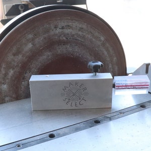 Pode incluir: Um close-up de um jig de lixar com um guia de acrílico transparente e uma base de metal. A base tem o texto "Maker Select" gravado. O jig está conectado a uma lixadeira com um disco de lixar marrom grande.