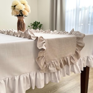 Puede incluir: Un mantel beige y blanco con volantes y un camino de mesa a juego. El camino de mesa está centrado en la mesa y tiene un borde con volantes. Un jarrón con flores color crema está sobre la mesa. El mantel tiene un borde con volantes blanco.