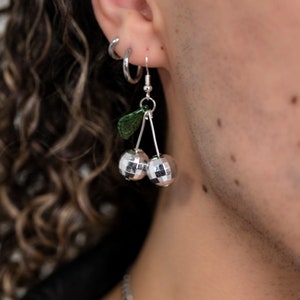 May include: A silver-toned dangle earring featuring two spherical disco ball charms arranged like cherries, suspended from a silver hook. A small, textured green leaf charm is attached to the top of the stems, adding a contrasting color element.