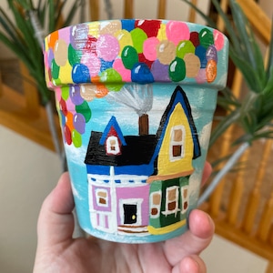 May include: A hand-painted terracotta flower pot with a scene of a house with balloons, inspired by the movie Up. The house is painted in shades of pink, yellow, green, and blue, with a black roof. The balloons are multi-colored and are floating above the house.