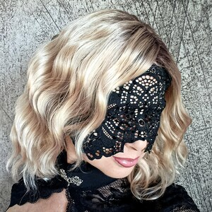 May include: A black lace eye mask with an intricate floral design. The mask covers the eyes and part of the nose. The person is wearing a black lace top with a decorative brooch. The hair is blonde with darker roots.