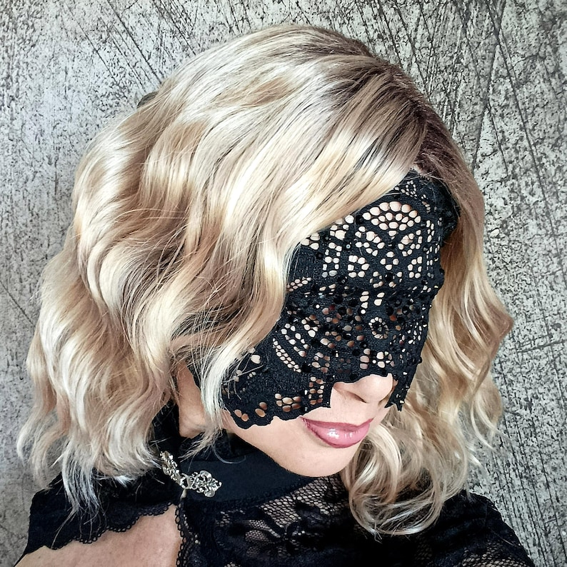 May include: A black lace eye mask with an intricate floral design. The mask covers the eyes and part of the nose. The person is wearing a black lace top with a decorative brooch. The hair is blonde with darker roots.
