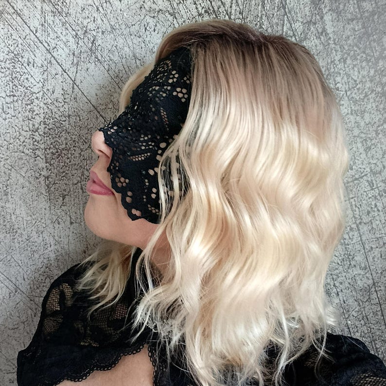 May include: Black lace eye mask with a floral pattern, covering the eyes and nose. The mask is worn by a person with wavy blonde hair. The background is a textured gray wall. The mask is part of a lingerie set.